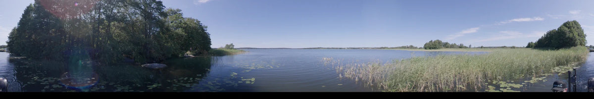 A daytime view from a boat on a lake in Upplands-Bro, Sweden, during summer. Clear skies overlook calm waters with lily pads and a forested shoreline.