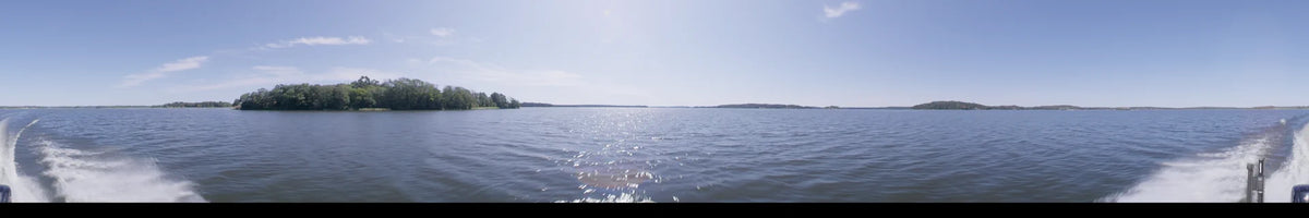 A daytime view from a boat in Upplands-Bro, Sweden, featuring glistening water and a distant forested shoreline under a clear summer sky.