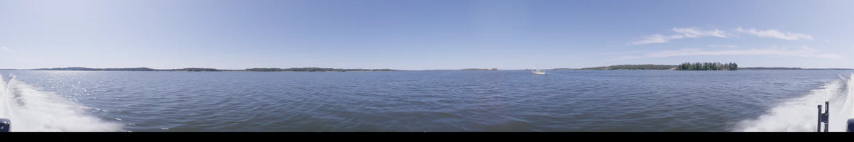 A distant, tree-lined coast borders the open water of Upplands-Bro, Sweden, seen from a boat under a clear summer sky during the day.