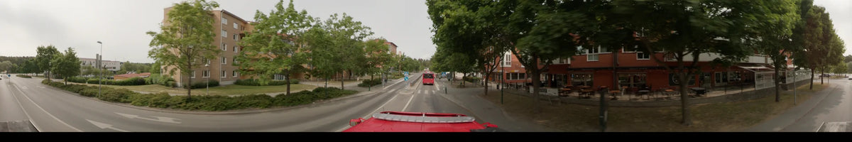 A bus follows another bus along a tree-lined road in Märsta, Sweden, on an overcast summer morning, passing a red building with a Subway sign.