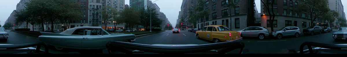 A car drives through a New York City street on an overcast spring morning, passing tall apartment buildings and yellow taxis parked along the curb.