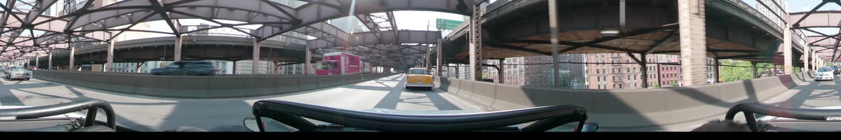 A yellow vintage car travels along the Long Island Expressway in New York, passing a pink truck under a steel overpass on a clear spring day.