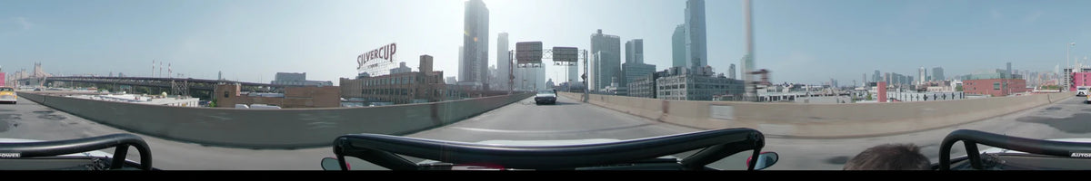 An urban skyline and overhead road signs frame a car traveling along a New York highway in the United States during a clear spring day.