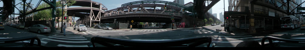 Beneath a steel overpass on Queens Plaza South in New York, United States, a car travels during a clear spring day, passing a cyclist and traffic barrels.