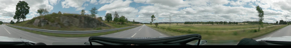 A multi-lane road between Upplands-Bro and Uppsala is flanked by open green fields and scattered trees under a partly cloudy sky.