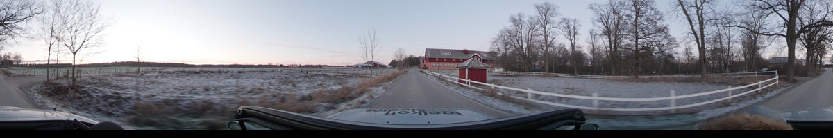 A car drives along a road in Lennartsnäs, Sweden, during a clear autumn afternoon, passing traditional red buildings and fields covered in light frost.
