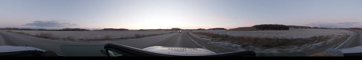 A car drives on a road in Lennartsnäs, Sweden, following another vehicle during a clear autumn afternoon. Open fields and distant trees line the horizon.
