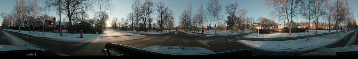 Passing a lone pedestrian and red buildings, a car drives along a road with bare trees and snow in Stockholm, Sweden, during a clear winter sunset.
