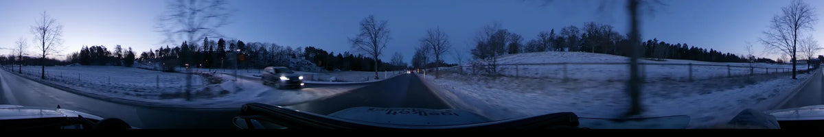 A car drives along a snowy road in Stockholm, Sweden, at dawn. Leafless trees and snow-covered fields line the clear winter landscape under a blue sky.