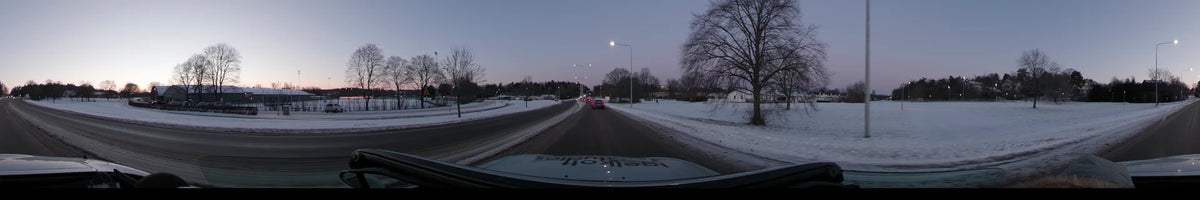 Streetlights line a snowy road in Stockholm, Sweden, during a clear winter dawn. A car follows traffic past leafless trees and snow-covered fields.