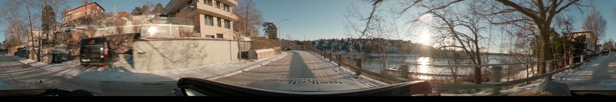Moving through Stockholm, Sweden, a car follows a snowy road lined by a waterfront railing and hillside houses during a clear winter sunset.