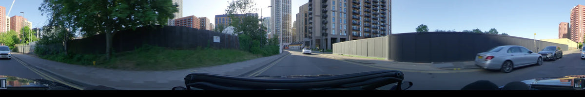 Contemporary apartment complexes and green foliage line the road as a car travels through London, United Kingdom, on a clear spring day.