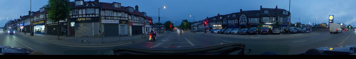 Local shops and a motorcyclist at red traffic lights line the road as a car drives through London, United Kingdom, on an overcast spring evening.