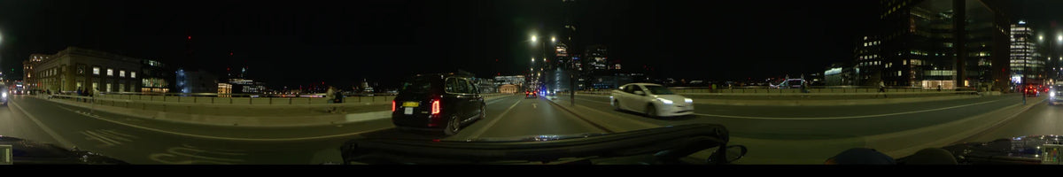 A black taxi and white car share the road during a clear spring night in London, United Kingdom, with illuminated modern buildings in the distance.