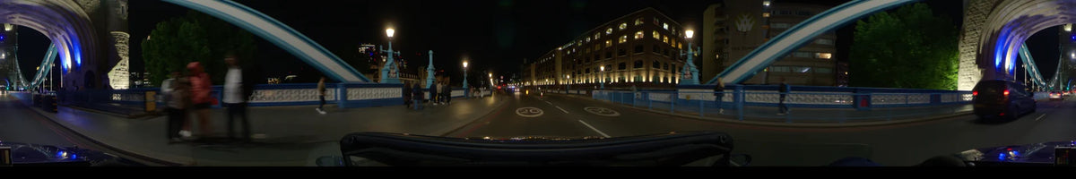 Pedestrians line the sidewalk while a car passes a large brick building with illuminated arched windows in London, United Kingdom, during a clear spring night.