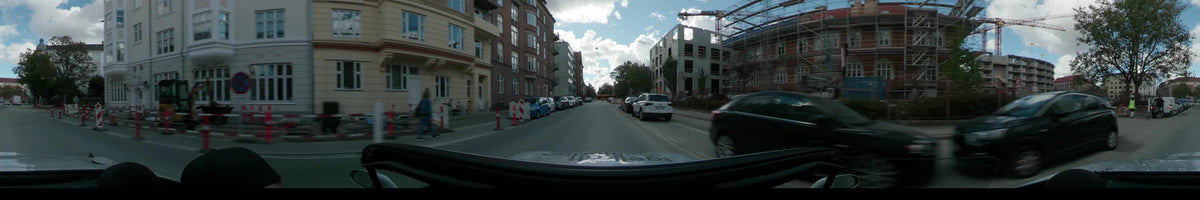 A city street in Copenhagen lined with traditional brick apartments and a modern concrete building under construction, with parked cars and road barriers along the way.