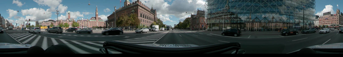 A wide city road in Copenhagen featuring ornate red brick buildings and a modern glass facade, with white vans and cars driving through moderate traffic.