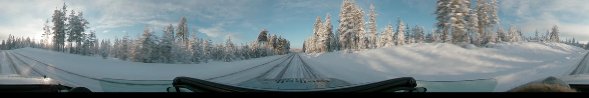 The snowy forests of Dalarna, Sweden, surround a straight road where a car travels during a partly cloudy winter day.