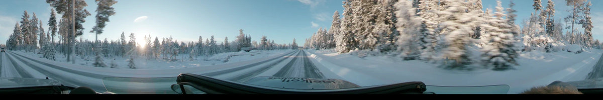 Snow-covered pine trees line a road in Dalarna, Sweden, as a car drives past a white trailer on a partly cloudy winter day.