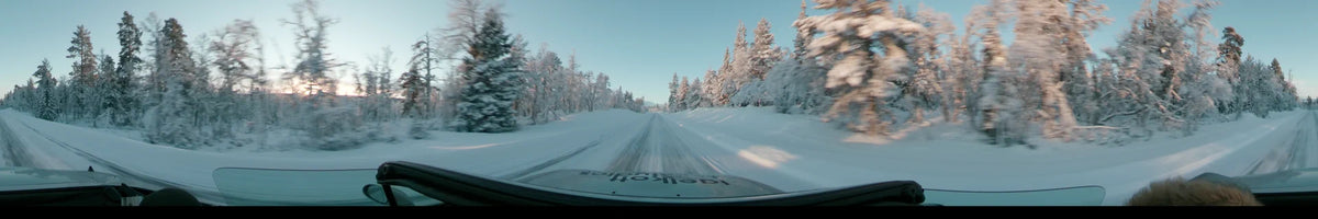 Snow-covered pines flank a white road in Grövelsjön, Sweden, as a car drives forward under a partly cloudy sky during a winter dusk.