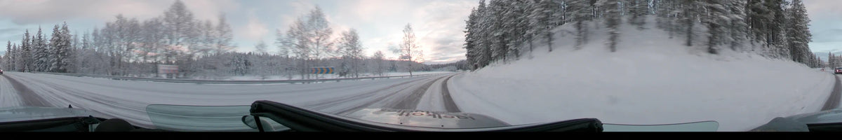 A car travels on a snow-covered road through Grövelsjön, Sweden, during a partly cloudy winter afternoon, with frost-laden trees lining the route.