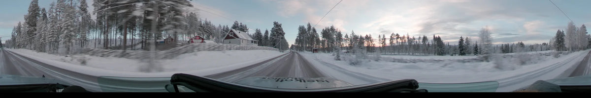 A white house and snow-covered pines line a road in Grövelsjön, Sweden, as a car drives through on a partly cloudy winter afternoon.