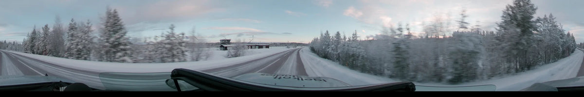 During a partly cloudy winter afternoon in Grövelsjön, Sweden, a car travels along a snowy road past buildings and snow-covered trees.