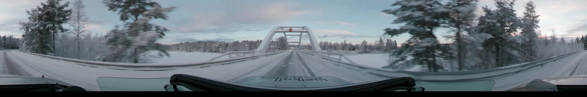 Crossing a white arch bridge in Idre, Sweden, a car travels on a snowy road during a partly cloudy winter afternoon amidst frosted trees.