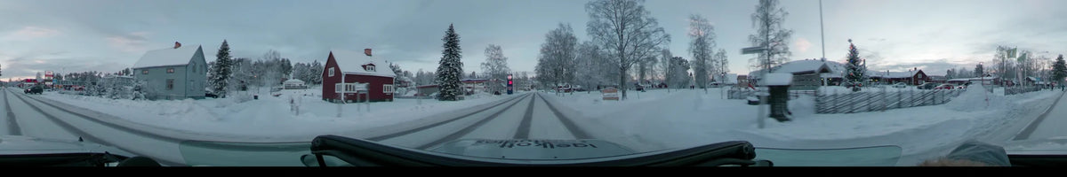 Passing through Idre, Sweden, a car moves along a snowy road bordered by buildings and frosted trees on a partly cloudy winter afternoon.