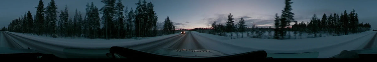 Frosted trees line a snowy road in Dalarna, Sweden, where a car passes another vehicle under a partly cloudy winter sunset sky.