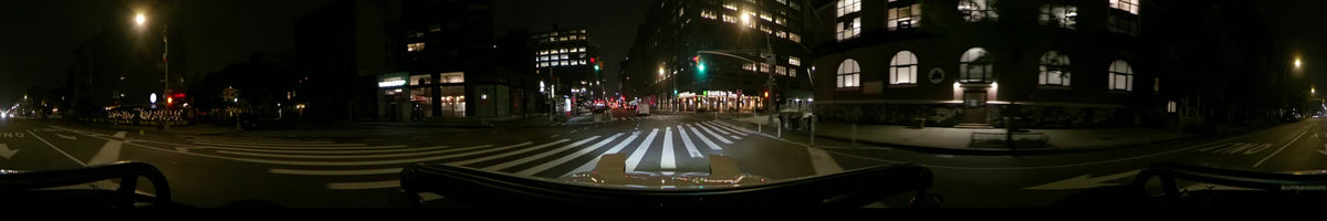Towering buildings and city lights frame a car passing a Shake Shack and a pedestrian crosswalk in New York at night during spring.