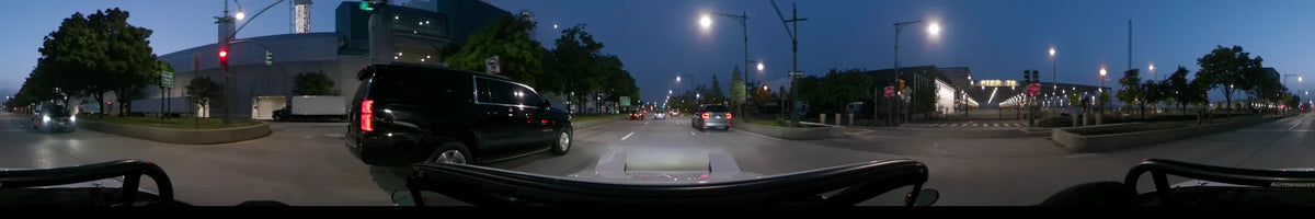 During a spring dawn in the United States, a car drives past a black SUV on a tree-lined New York street under glowing streetlights.