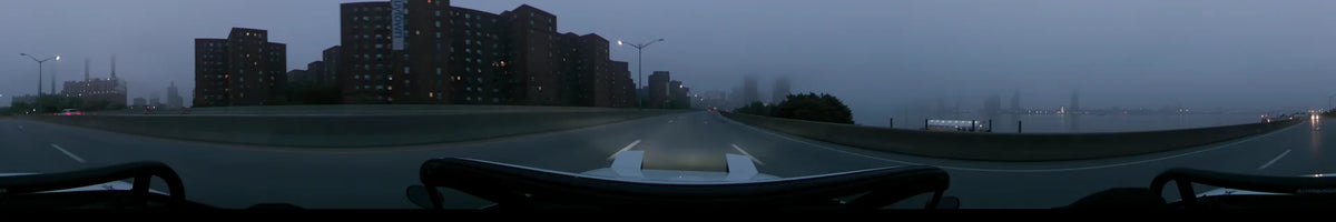 Large brick buildings and spring greenery flank a New York road as a car proceeds through the fog at dawn in the United States.