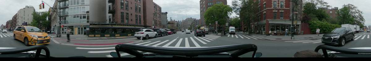 Green trees and brick buildings frame a New York street as a car crosses a white pedestrian crossing on an overcast spring day.