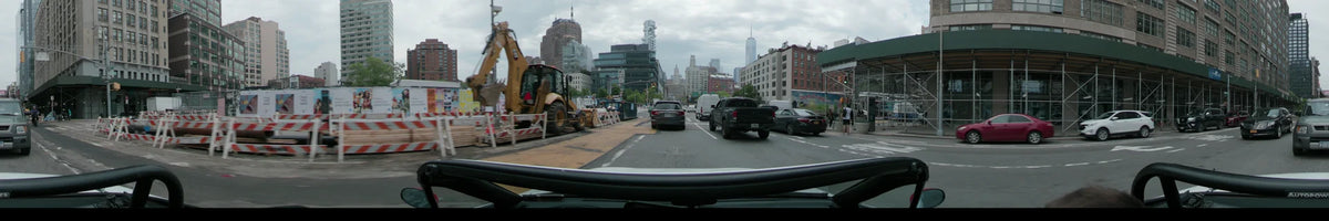 One World Trade Center is visible in the distance as a car navigates New York traffic and a roadside construction zone on an overcast spring day.