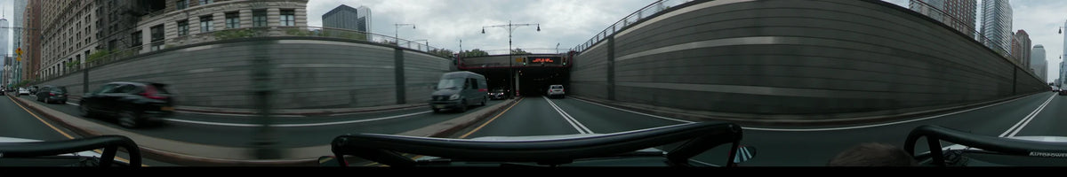 Flanked by concrete walls, a car approaches a New York tunnel on an overcast spring day, following a white SUV and passing a grey van.