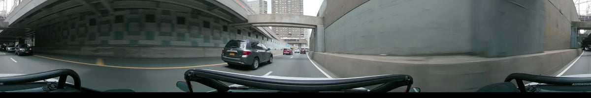 Tall brick buildings tower over a New York road on a partly cloudy spring day, where a car follows a red vehicle past concrete walls.