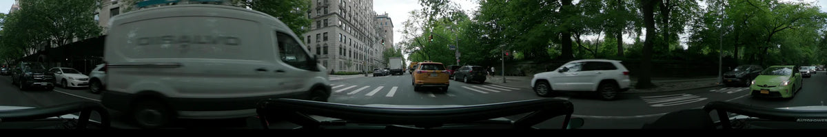 On a partly cloudy spring day, a car follows a yellow vehicle and white truck through New York, passing by tall residential buildings and green trees.