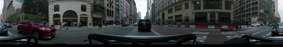 A black SUV leads a car past pedestrians and tall urban buildings on a New York street during a partly cloudy spring day.