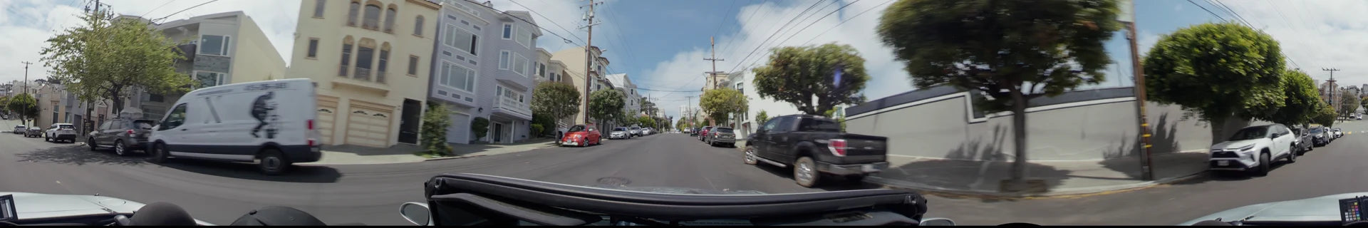 A residential street in San Francisco lined with classic multi-story houses, leafy green trees, and parked cars under a partly cloudy sky.