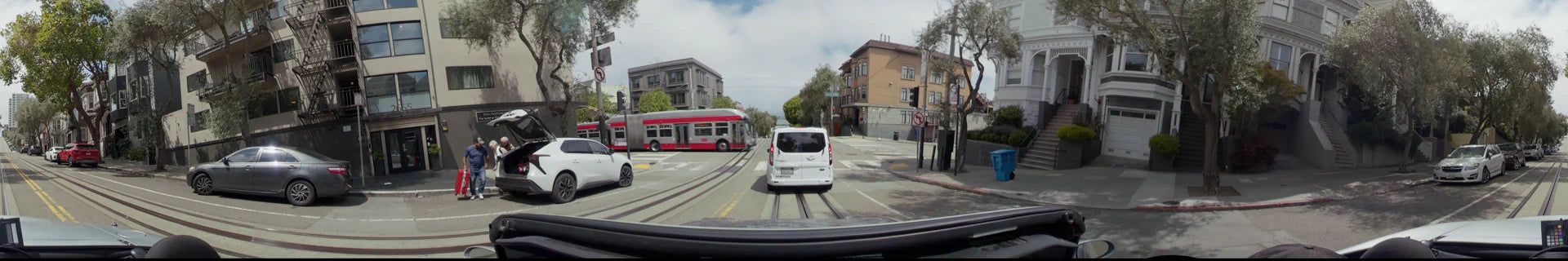 A city street in San Francisco with a white van and a red bus crossing a track-lined road surrounded by residential buildings.