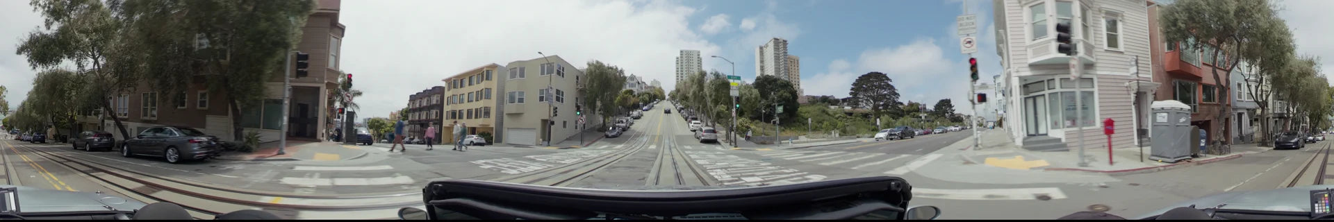 A city street in San Francisco with tram tracks, parked cars, and high-rise residential buildings under a partly cloudy sky.