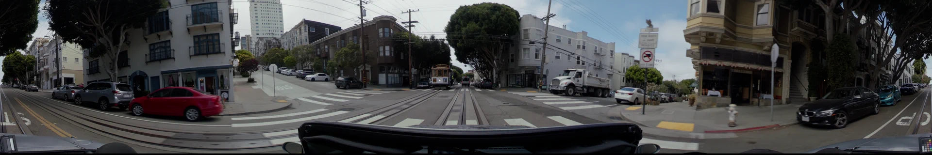 A San Francisco street featuring a cable car and a white truck crossing a track-lined road surrounded by residential buildings and lush green trees.