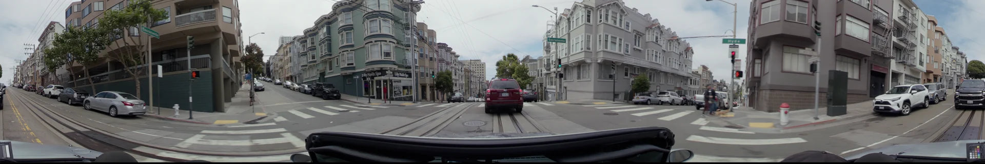 A red SUV drives along Sacramento Street in San Francisco, passing multi-story residential buildings and tram tracks under a partly cloudy sky.