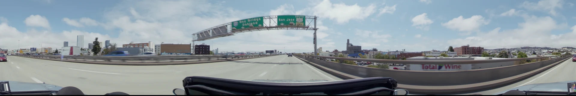 A multi-lane highway in San Francisco with overhead signs for the Bay Bridge and San Jose, featuring light traffic and city buildings under a partly cloudy sky.