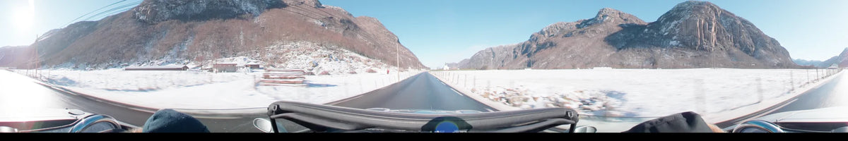 View from a car driving on a road in Stavanger, Norway, during a clear spring day. Snowy landscapes and mountains flank the way, with a small white building ahead.
