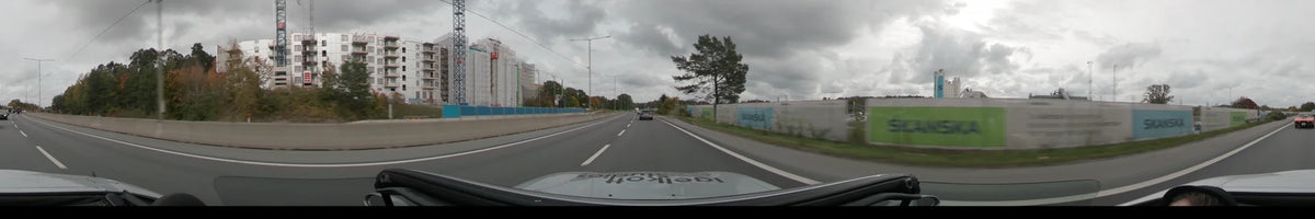 Construction fences and autumn trees border a multi-lane road in Stockholm, Sweden. The car travels under an overcast sky during the day.
