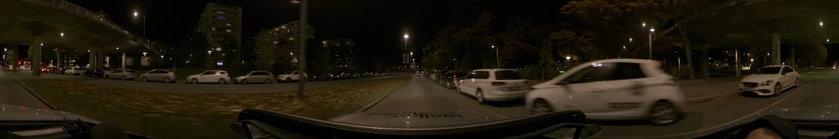 Under a clear night sky in Stockholm, Sweden, a car passes by autumn trees, parked vehicles, and buildings illuminated by streetlights.