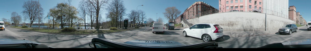 A car drives through Stockholm, Sweden, on a clear spring day. The view captures white vans on a city street bordered by bare trees and buildings.