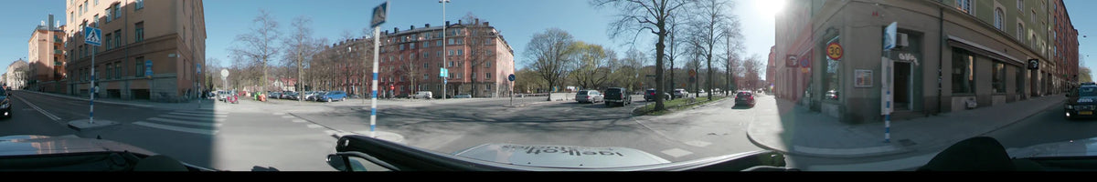 Past a pink apartment building and budding trees, several cars travel along a Stockholm, Sweden city street during a clear spring day, seen from a car.